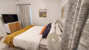 A bedroom with a bed, a TV on a stand, a painting on the wall, and curtains.