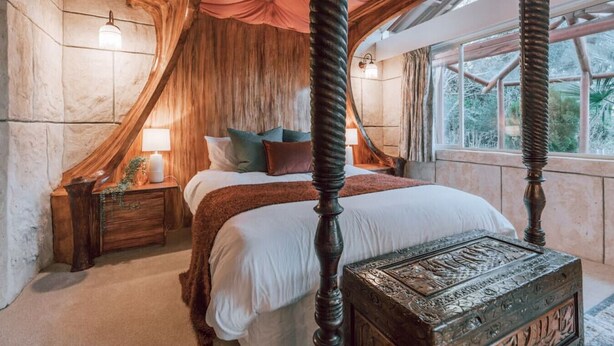 A four-poster bed with a canopy, a nightstand with a lamp, and a decorative chest in a room with stone walls and a large window.