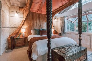 A four-poster bed with a canopy, a nightstand with a lamp, and a decorative chest in a room with stone walls and a large window.