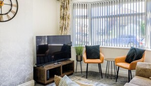 Ein Wohnzimmer mit einem Flachbildfernseher, zwei orangefarbenen Sesseln, einem kleinen runden Tisch und einem Fenster mit Jalousien.