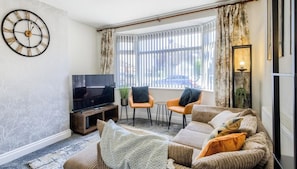 A living room with a flat-screen TV, two armchairs, a sofa, a clock, and a window with curtains. - 52NRC Unli Wi-fi 3 Bedroom House (Coventry)