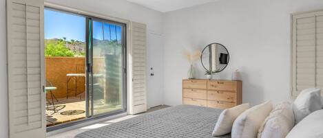 Een slaapkamer met een groot bed, een dressoir, een spiegel en een schuifdeur die leidt naar een terras met tuinmeubilair.