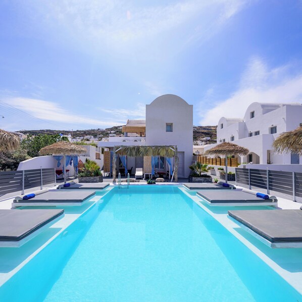 A pool area with a clear blue pool, sun loungers, and a view of white buildings with a thatched roof structure.