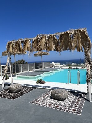 A rooftop pool area with a clear view of the ocean, lounge chairs, and a pergola with dried palm leaves.