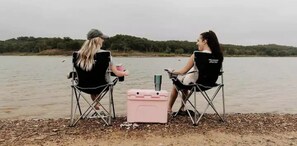 Zwei Personen sitzen auf Klappstühlen am Seeufer, dazwischen ein Kühlbox und eine Wasserflasche.