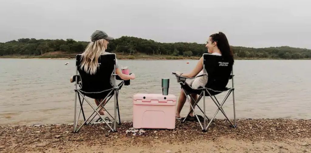 Deux personnes sont assises sur des chaises pliantes, près d’un lac, avec une glacière et une bouteille d’eau posées entre elles.