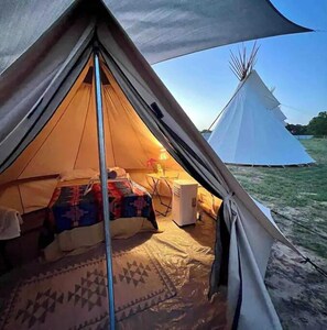 Une tente équipée d’un lit, d’une petite table et d’un chauffage. On aperçoit un tipi blanc à l’arrière-plan.