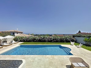Une piscine avec des chaises longues, une vue sur la mer et un jardin agrémenté de plantes en fleurs.