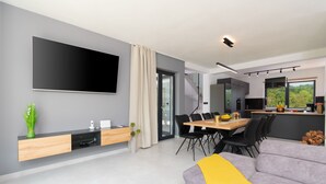 Una sala de estar moderna con un televisor de pantalla plana, un comedor con una mesa y sillas de madera, y una cocina con un ventanal grande.
