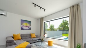 A modern living room with a grey sofa, yellow cushions, a coffee table, and a large window with a view of a swimming pool and greenery.