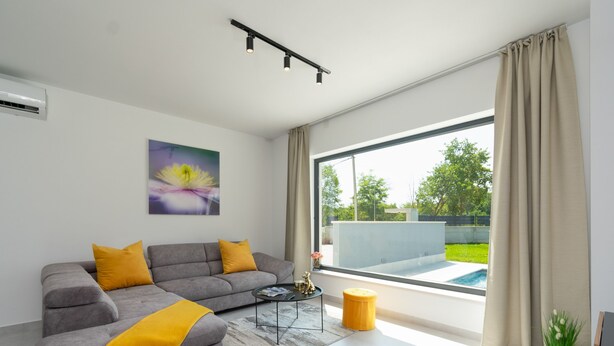 Una sala de estar moderna con un sofá gris, cojines amarillos, una mesa de centro y un amplio ventanal con vistas a una piscina y follaje.