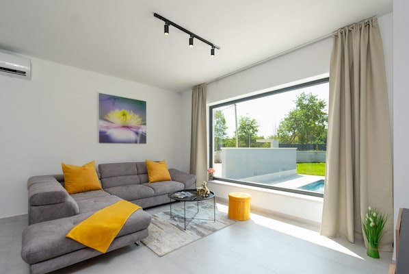 A modern living room with a grey sofa, yellow cushions, a coffee table, and a large window with a view of a swimming pool and greenery.