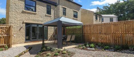 A modern two-story house with a stone facade, a covered patio with outdoor furniture, and a wooden fence.