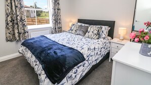 Une chambre à coucher avec un lit, une table de chevet, une lampe, une commode et une fenêtre avec des rideaux.