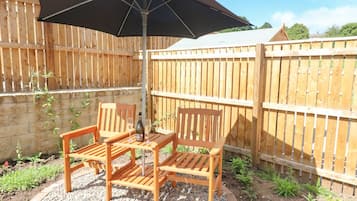 Un patio de madera con dos sillas y una mesa bajo un paraguas negro, rodeado por una valla de madera y un muro de piedra.