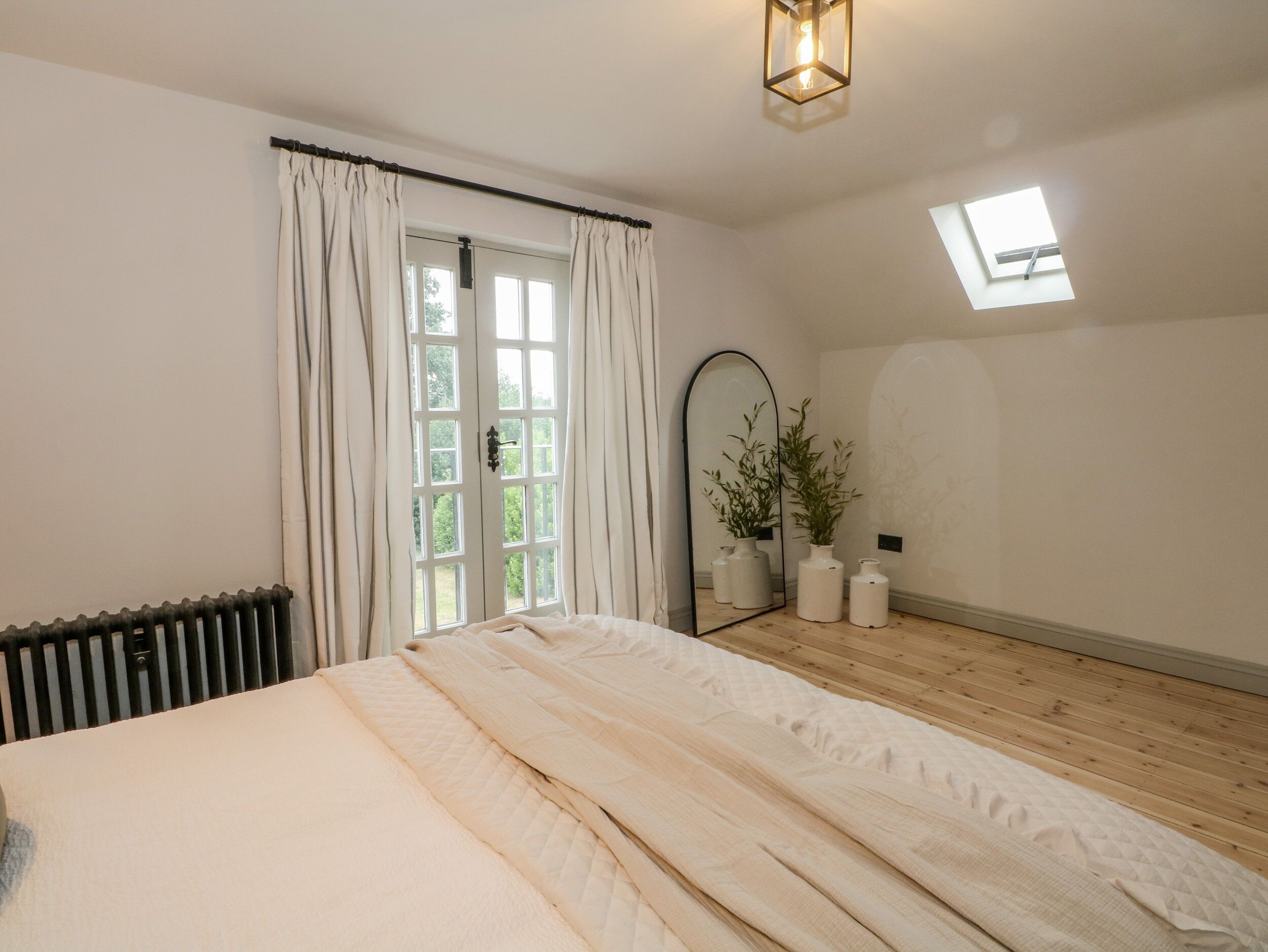 A bedroom with a bed, a radiator, a window with curtains, a mirror, and a skylight.
