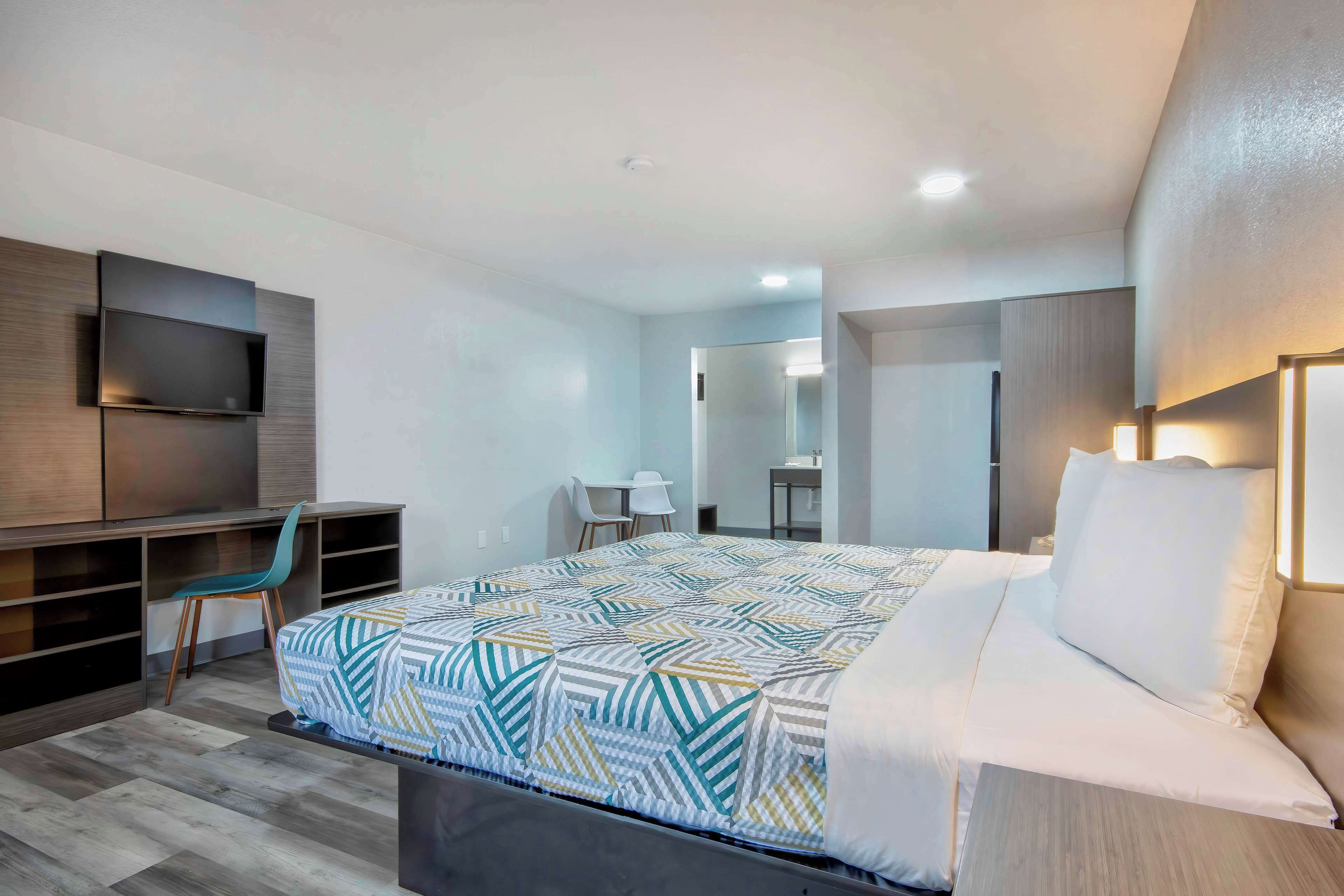 A modern hotel room with a large bed, a desk, a TV, and a bathroom visible in the background.