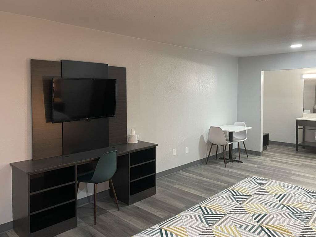A modern hotel room with a flat-screen TV mounted on the wall, a desk with a chair, a small table with a chair, and a patterned bedspread.
