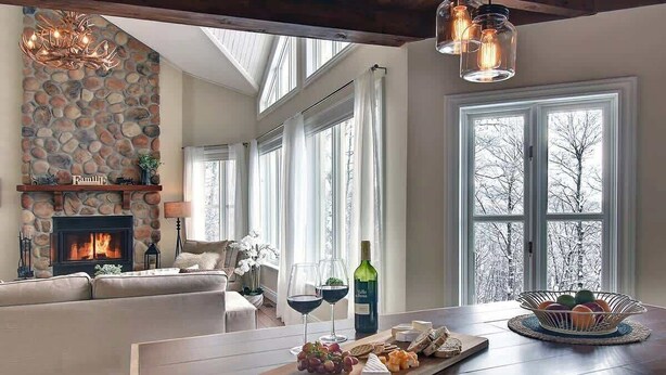 Een gezellige woonkamer met een stenen openhaard, een houten eettafel met kaas en druiven, en uitzicht op bomen door grote ramen.