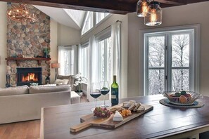 A cozy living room with a stone fireplace, a wooden dining table with cheese and grapes, and a view of trees through large windows.
