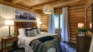 Een gezellig houten blokhut slaapkamer met een houten plafond, een groot bed met veel kussens, een hanglamp en een raam met gordijnen.