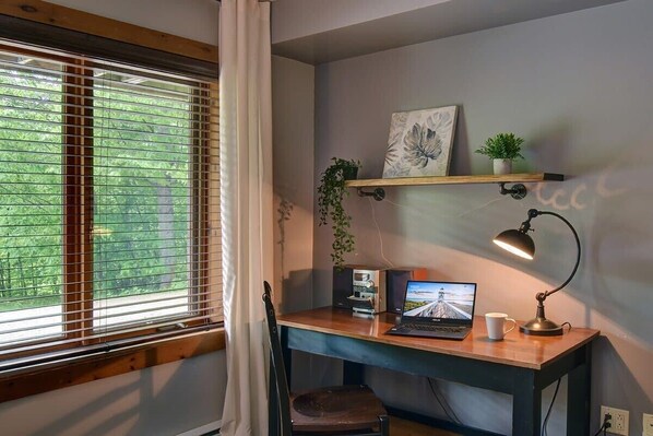 A room with a wooden desk, a laptop, a desk lamp, a chair, a window with blinds, a shelf with a plant and a picture, and a wall-mounted power outlet. - Chic Hot tub in mountain Tremblant Area (Labelle)