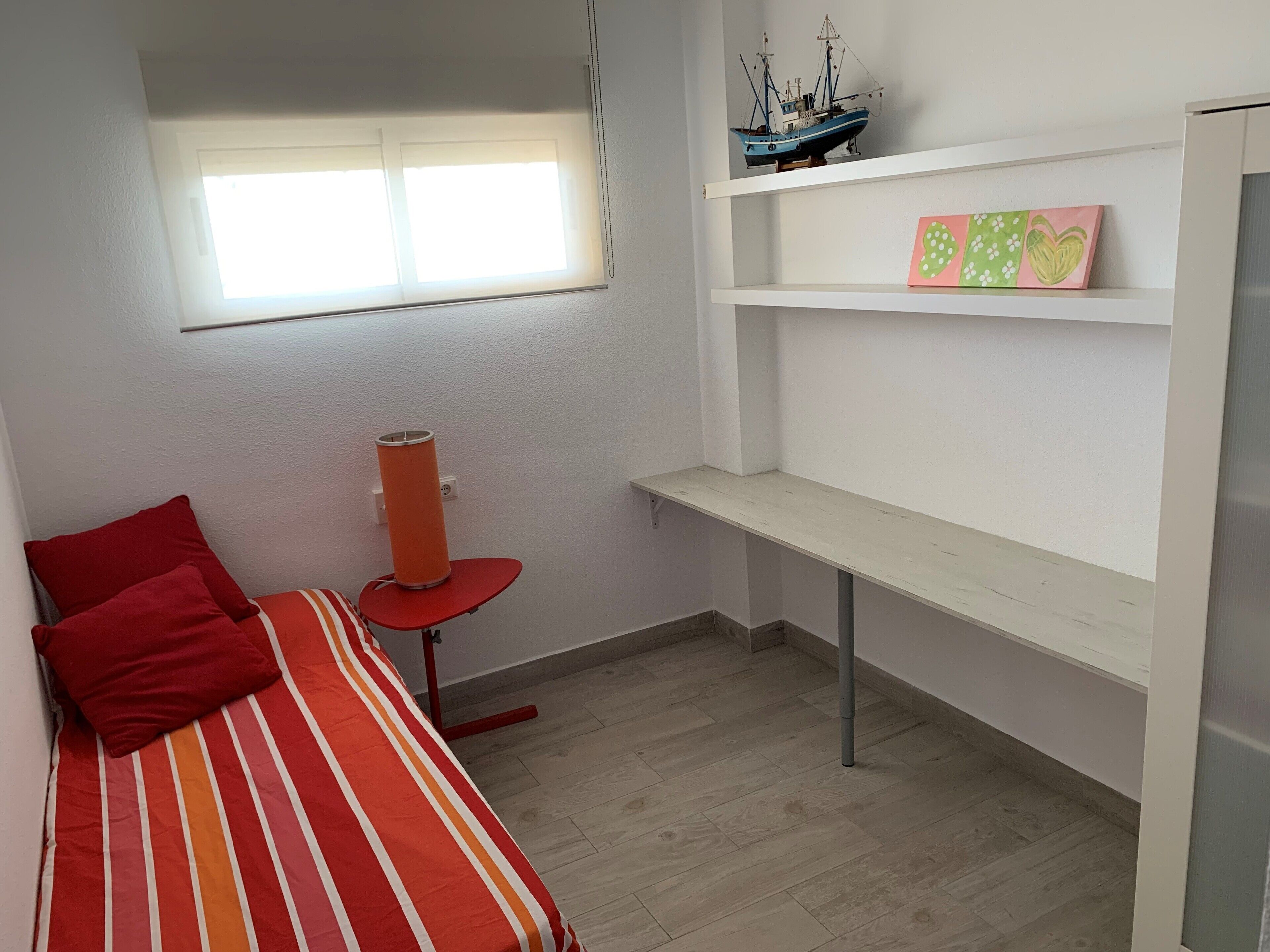 Een kleine, netjes ingerichte kamer met een bed, een bureau, een plank met decoratieve objecten en een raam.