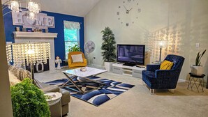 Een woonkamer met een open haard, een televisie, een gele fauteuil, een blauwe fauteuil en een kroonluchter.
