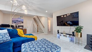 Een moderne woonkamer met een blauwe bank, een gestreept voetenbankje, een flatscreen televisie en een trap naar een hoger gelegen verdieping.