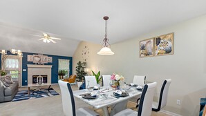Een eetruimte met een tafel gedekt voor zes personen, omringd door witte stoelen met zwarte accenten. Daarboven hangt een kroonluchter, en er zijn twee aan de muur bevestigde schilderijen.