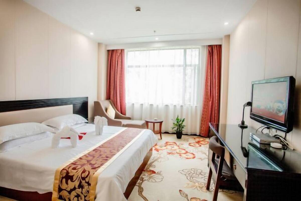 Habitación de hotel con cama, una silla, una mesita, televisión y ventana con cortinas.