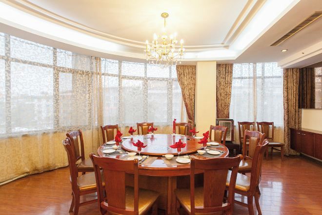 A dining room with a round table set for six, surrounded by wooden chairs, large windows with curtains, and a chandelier.