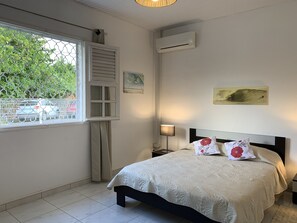 Un dormitorio con cama, mesita de noche, lámpara, aire acondicionado y ventana con vistas a árboles y coches estacionados.