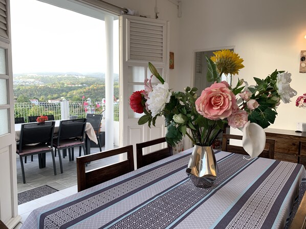 A dining area with a table set for two, a vase with flowers, and a view of a balcony with chairs and a railing.