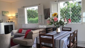 A living room with a sofa, dining table, and chairs. There is a fireplace and a window with shutters. - Magnificent villa with free parking ideally located. (Schoelcher)