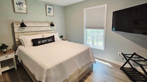 Een slaapkamer met een houten hoofdeinde, een bed met wit beddengoed, twee ingelijste bloemen afbeeldingen, een raam met zonwering, en een aan de muur bevestigde televisie.