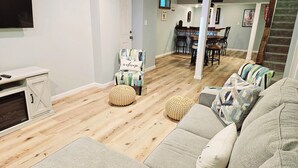 Een woonkamer met een bank, poef en een stoel met een kussen waarop 'welkom' staat. Achtergrond toont een eethoek met een tafel en stoelen.