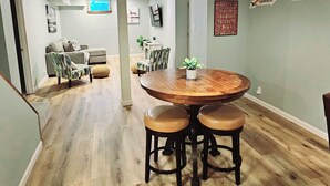 Een eethoek met een ronde houten tafel en drie krukken, een hangende hanglamp en een aan de muur bevestigde televisie.
