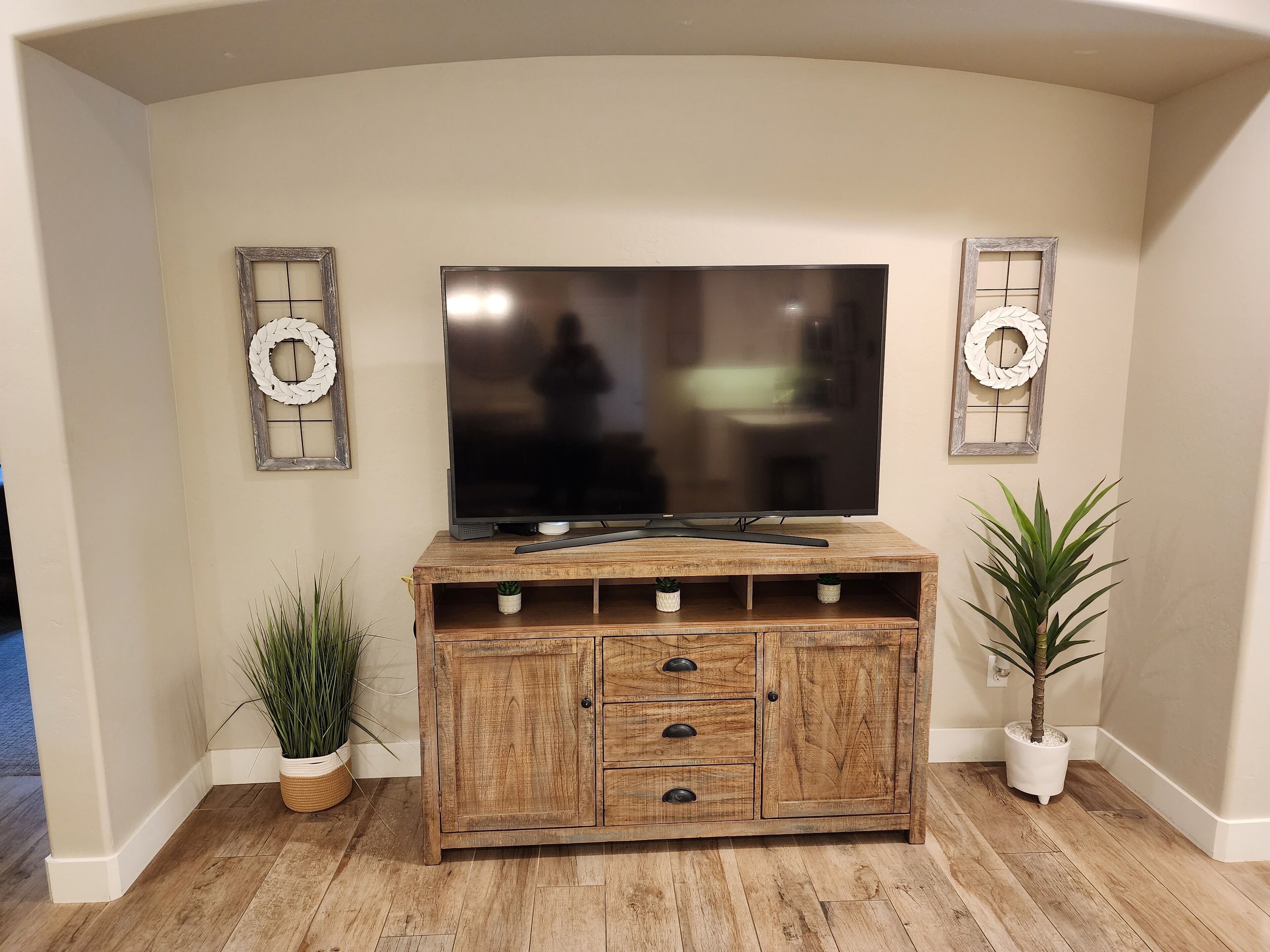 A wooden entertainment center with a flat-screen TV, two decorative wreaths, and potted plants.