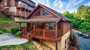 A wooden cabin with a balcony, a stone foundation, and a parking area.