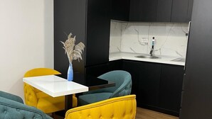 Een compacte keuken met een wit aanrecht, zwarte kastjes en een klein spoelbakje. Er staan twee stoelen, een geel en een teal, en een vaas met gedroogde planten op het aanrecht.