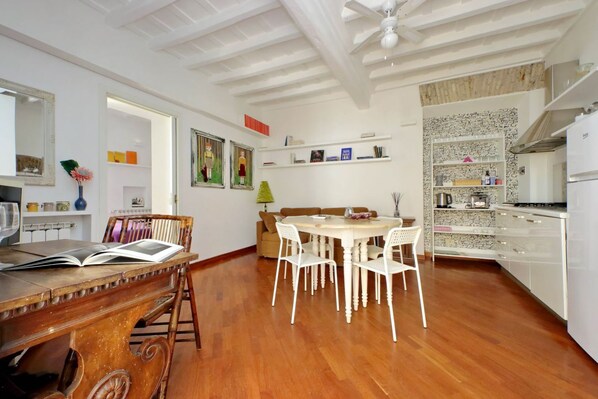 A dining area with a wooden table and chairs, a ceiling fan, and a kitchen area with white cabinets and a stone backsplash.