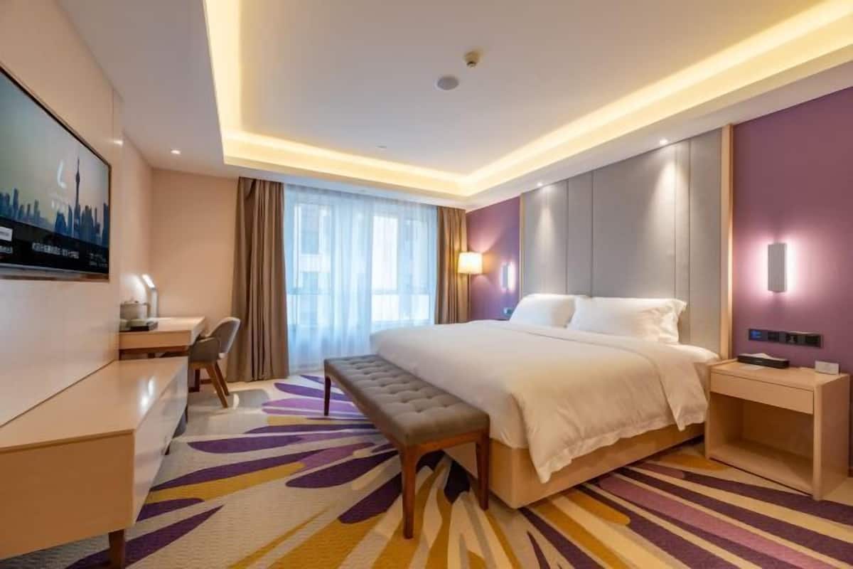 A hotel room with a bed, a desk, a bench, a TV, and a window with curtains.
