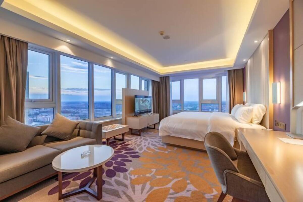 A hotel room with a large bed, a sofa, a desk, a TV, and a view of the city.