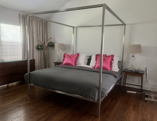 Un lit à baldaquin avec une couvre-lit grise et des coussins roses, une tête de lit en bois, une table de chevet avec une lampe et une fenêtre avec des rideaux.