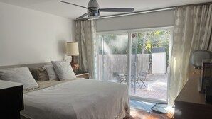 Un dormitorio con cama, ventilador de techo, mesita de noche con lámpara y vista al patio trasero a través de una puerta corrediza de vidrio.