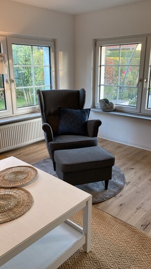 A modern living room with a grey sofa, a white coffee table, and a round rug. There are two large windows providing views of a garden.