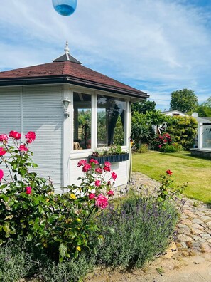 A white house with a covered porch, a garden with pink roses, and a lawn. - Thatched-roof vacation home with a view of the water, a garden, and a fireplace (Stahlbrode)