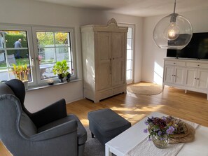 A living room with a sofa, armchair, TV stand, and a large window with plants. - Thatched-roof vacation home with a view of the water, a garden, and a fireplace (Stahlbrode)