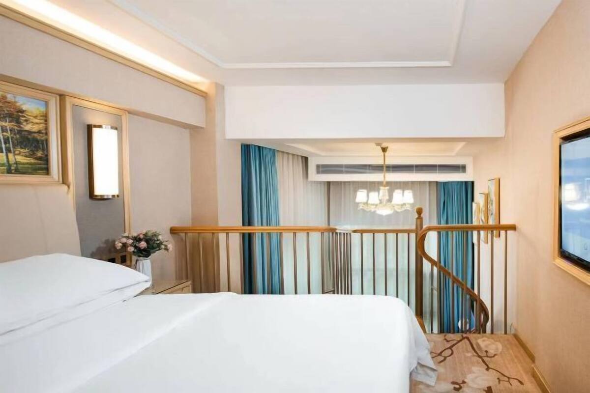 A hotel room with a bed, a nightstand, a vase with flowers, a chandelier, and a view of the outdoors through a window.